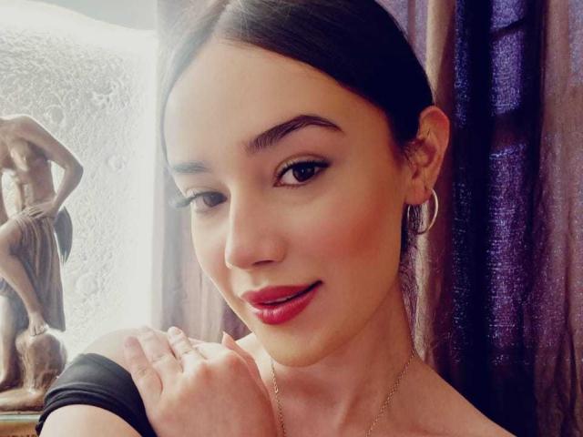 MariaLoft with dark hair, red lipstick, hoop earrings, and a black bracelet, smiling softly, holding her chin, standing next to a statue.