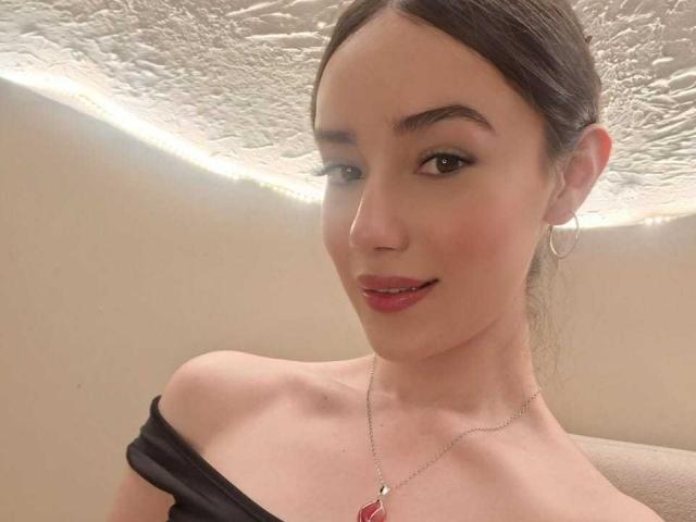 MariaLoft hat helle Haut, braune Augen und roten Lippenstift. Sie trägt ein schwarzes Oberteil mit einem Schulterauschnitt und eine rote Anhänger-Kette, Creolen und hat ein leichtes Lächeln..