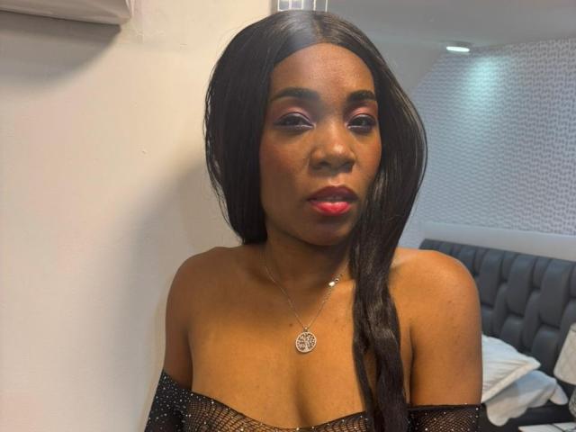 HilaryJaz with dark skin, long black hair, red lipstick, purple eyeshadow, black off-shoulder top, small silver necklace with circular pendant, looking at camera, in bedroom.
