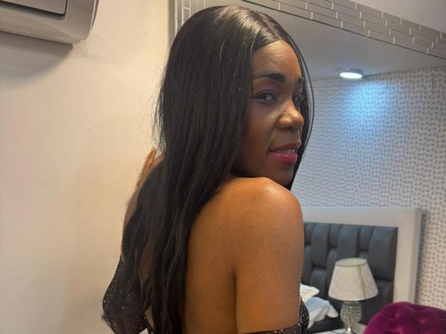 HilaryJaz with long black hair, looking over her shoulder, smiling, showing her back with sparkly black top, red lipstick, and dark skin, in a dimly lit room.