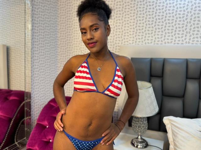 MartinaRoss with dark skin, short curly hair, wearing a red and white striped bikini top and blue polka dot bottoms, hands on hips, smiling, standing in a bedroom.