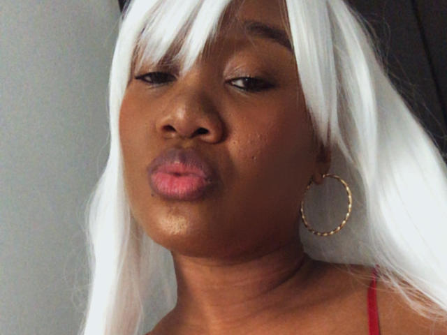 Close-up of AshantyHotLove with dark brown skin and white straight hair. She has full pink lips, slightly parted, and is wearing a red strap. Her right ear has a large gold hoop earring. Her eyebrows are thin and she has dark eyes with a subtle eyeliner. The hair has a straight texture, covering parts of her forehead. She is looking to the left. The lighting is even, highlighting her smooth skin texture.