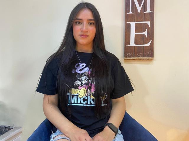 SaraThoms with long black hair, wearing a black "Mickey's" t-shirt and blue jeans, sitting on a chair, has a black watch on her left wrist, smiling.