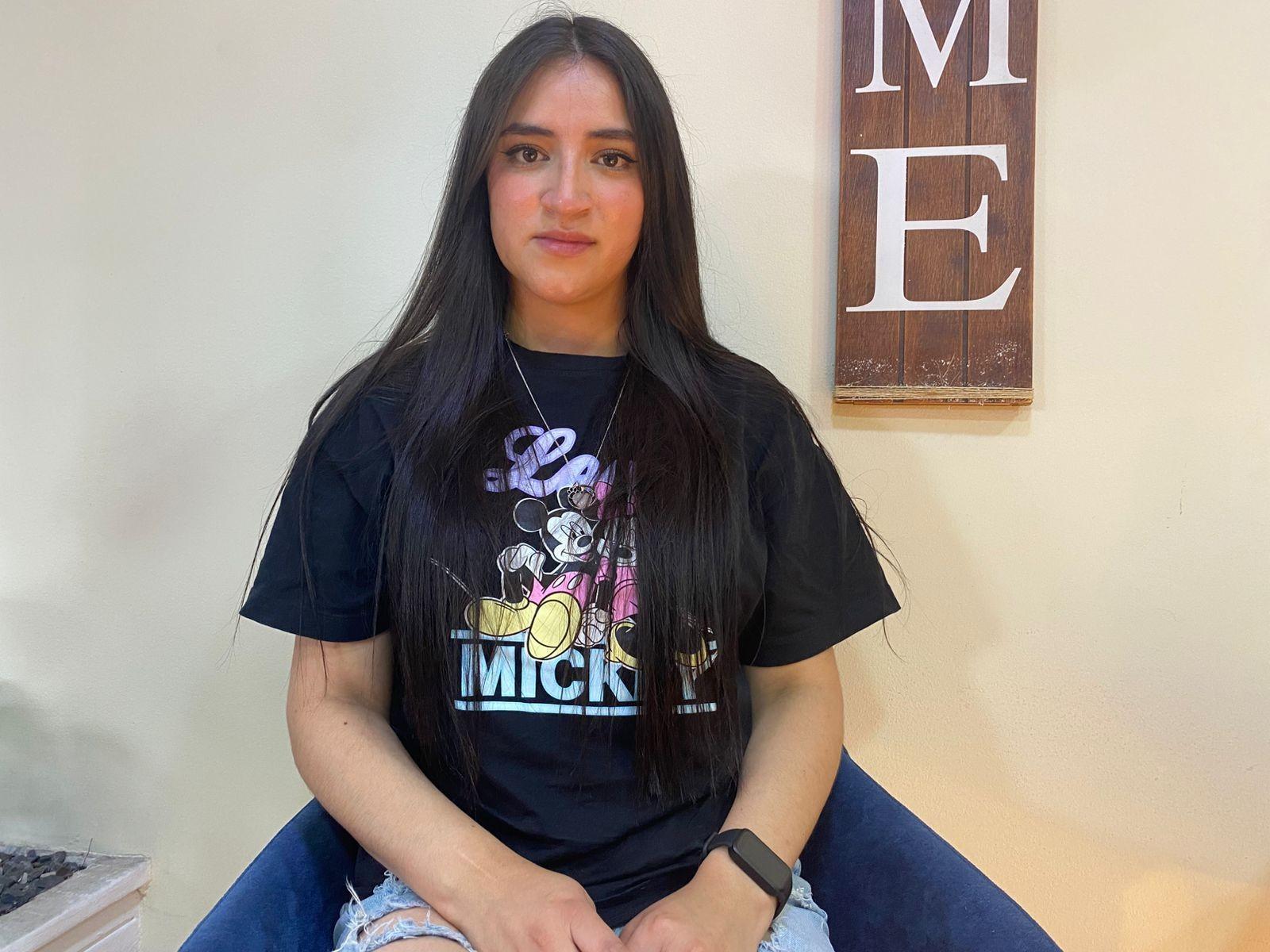 SaraThoms with long black hair, wearing a black "Mickey's" t-shirt and blue jeans, sitting on a chair, has a black watch on her left wrist, smiling.