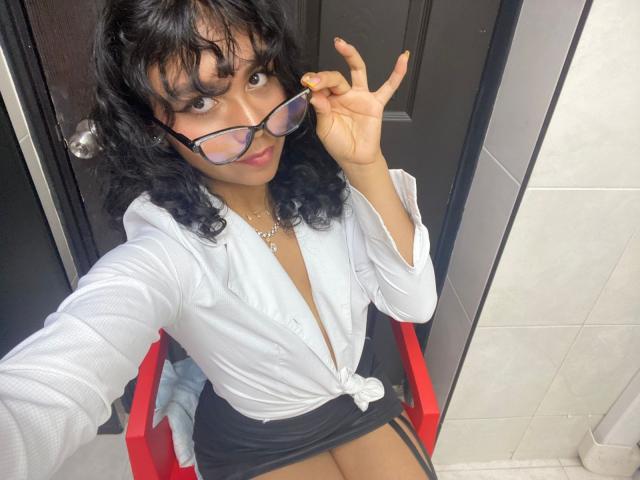Julia Bennett, met krullend zwart haar, draagt een wit overhemd met knoopjes, een zwarte rok en een bril. Ze maakt een vrede-teken, toont haar gele nagels, en haar kleine borsten zijn zichtbaar..
