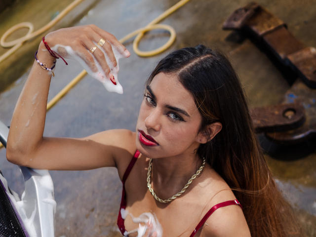Vanelope Russo, com cabelos castanhos longos, batom vermelho e sutiã vermelho, segurando um sabonete na mão, olhando para cima, usando uma corrente e pulseira douradas, em um ambiente metálico e úmido..