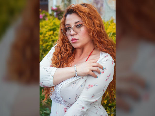 RosseHot69 with curly red hair, glasses, white floral dress, crossed arms, pink lipstick, bracelet, and serious expression, standing in a blurred garden. Boobs visible under dress.