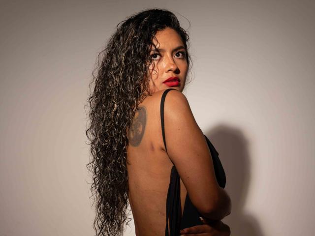 JedaZore mit lockigem, schwarzem Haar, rotem Lippenstift, einem Tattoo auf dem Rücken und einem schwarzen Kleid, die über die Schulter schaut und dabei ihren nackten Rücken und die schwarzen Träger zeigt. Die Brüste sind von der Seite sichtbar..