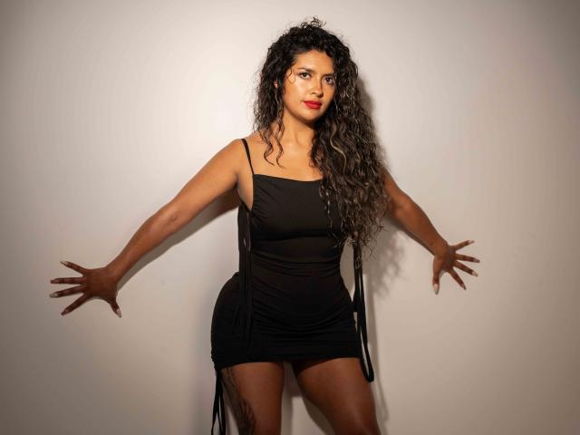 JedaZore, avec ses cheveux noirs bouclés, son rouge à lèvres rouge et sa robe noire transparente, les bras ouverts, se tient contre un mur blanc, mettant en valeur ses fesses et le tatouage sur sa cuisse droite..
