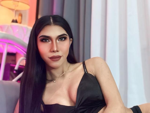 ZephyrLopez, avec ses longs cheveux noirs, portant un soutien-gorge en satin noir, montrant ses seins, a un joli visage avec du rouge à lèvres foncé et un collier, dans une pièce légèrement éclairée..