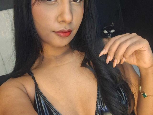 MariamLara with black hair, red lipstick, and a shiny black bikini top, showing off her tits and manicured nails, wearing a green ring, with a black cat in background.
