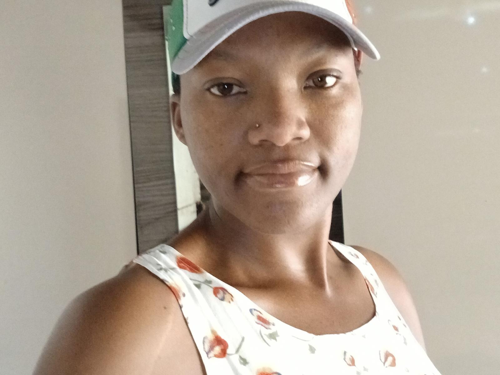 Bronwie with dark skin, small nose piercing, smiling, wearing a white cap and floral tank top, showing her shiny teeth. Her brown eyes are slightly upturned.