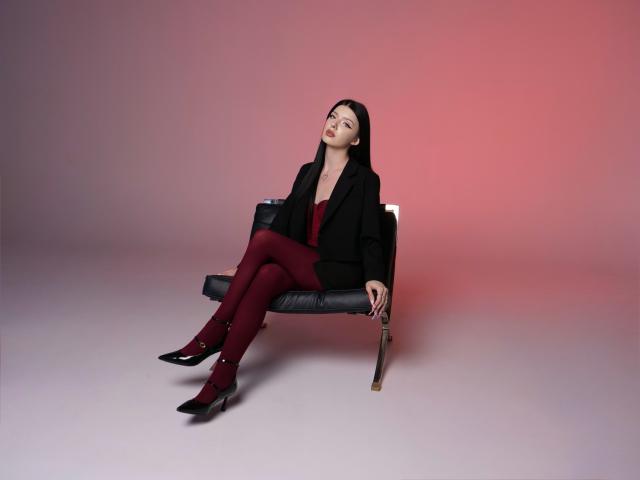 RoxyHopper, avec de longs cheveux noirs, un blazer noir, une robe rouge, des collants rouges, des chaussures noires, assise sur un banc en cuir noir, tenant un parapluie noir, la tête inclinée vers le haut, l'air détendue..