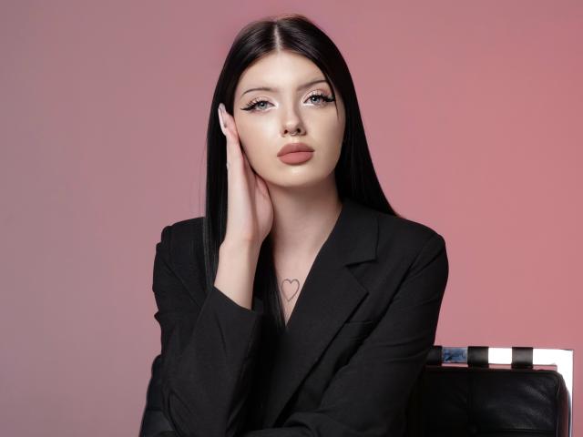 RoxyHopper, avec de longs cheveux noirs, portant un blazer noir, une main sur sa joue, un tatouage de cœur sur la poitrine, regardant l'appareil photo avec de pleines lèvres roses. Arrière-plan avec un dégradé rose..