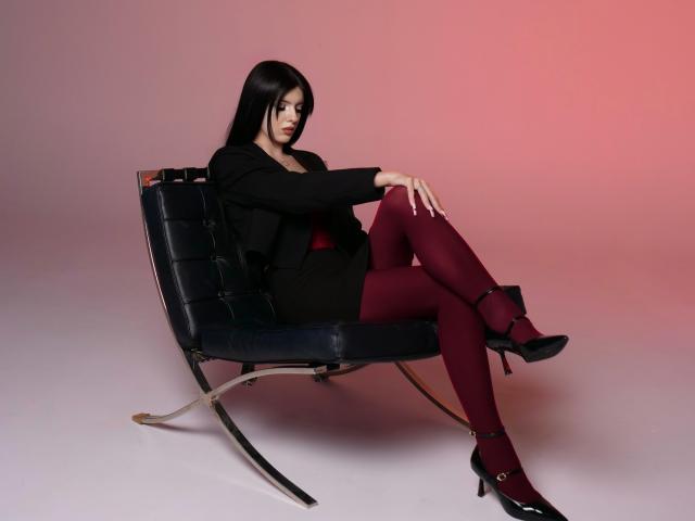 RoxyHopper sits on a black leather chair, wearing a black blazer, red dress, and maroon tights, with black high heels, legs crossed, and a serious expression.