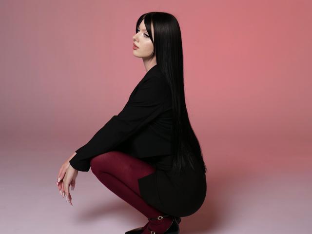 RoxyHopper, squatting, with long black hair, wearing a black jacket, red tights, and black shoes, looking to the side, showing her ass and small, cute tits.