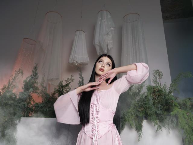 RoxyHopper with long black hair, pale pink dress, lace-up bodice, puffy sleeves, hands near face, greenery, white hanging garments, soft mist, looking up with a gentle expression.