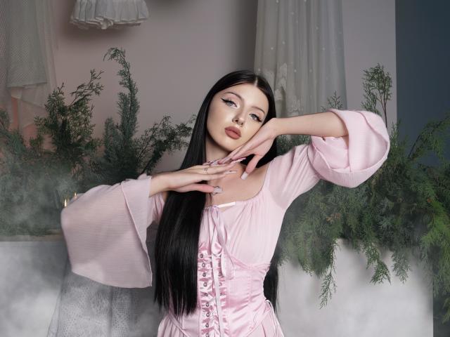 RoxyHopper with long black hair, pink dress, and laced front, posing with hands near face, showing off her pink-nail'd fingers, and her cute, small tits.