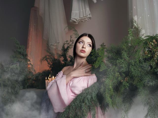 RoxyHopper stands amid green pine branches, her fair skin contrasting with the dark green foliage. She wears a light pink off-shoulder top, her right hand touching her neck. Her brown hair is straight and parted in the middle. She gazes slightly to the right, her expression neutral. Soft, warm light illuminates her from the left, highlighting her delicate features. Candles are partially visible among the pine branches, adding a gentle glow. The overall composition is balanced, with RoxyHopper centered and the pine branches framing her.