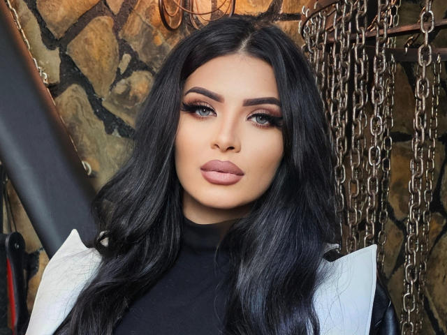 Devasha has long, black, wavy hair, blue eyes with thick, dark eyebrows, and full, pink lips. She wears a black turtleneck and a white, textured jacket. Her makeup is bold, with dark eyeshadow and eyeliner. The image has a realistic style with sharp details and a neutral, stone-patterned background. Chains hang from the right side, adding a metallic texture. Devasha's expression is neutral, and she is centered in the frame.