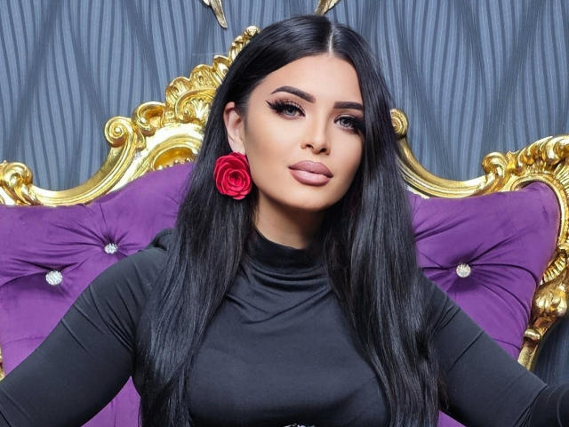 Devasha has long, straight black hair, full eyebrows, and thick eyeliner. She wears a black long-sleeve top and a red rose earring in her left ear. She sits on a purple, tufted velvet chair with a gold, ornate frame. Her lips are full and painted in a muted pink. The background has a striped, dark grey pattern. The gold frame has intricate carvings. The image is sharply focused, highlighting Devasha's facial features and the texture of the velvet.