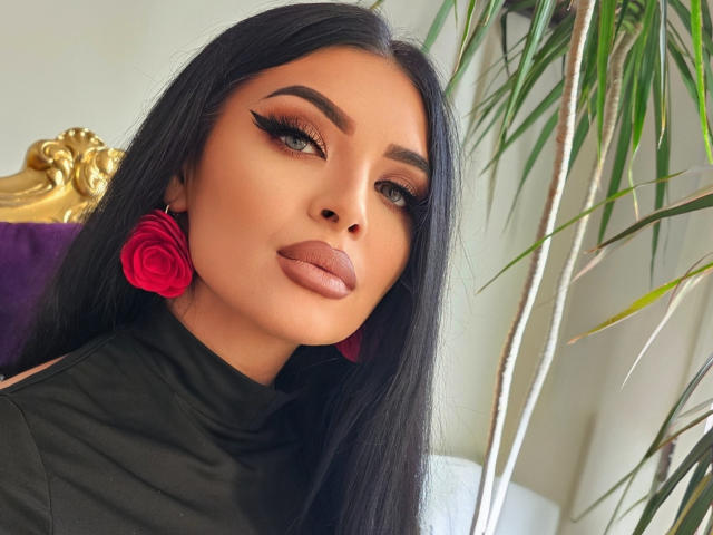 Devasha has long black hair, thick eyebrows, and wears red rose earrings with a black turtleneck. She's got full, glossy pink lips and a sexy, confident look.