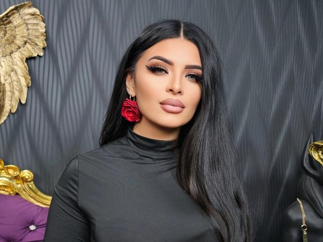 Devasha, a woman with long, straight black hair, wears a black turtleneck and a red rose earring in her right ear. Her makeup is bold, with thick, arched eyebrows, dark eyeliner, and pink lipstick. The background features a gold winged decoration on the left and a purple, tufted pillow on the bottom left. Her expression is neutral, and the texture of the background is a dark, patterned fabric. The image is sharply focused, with a clear, high-contrast composition.