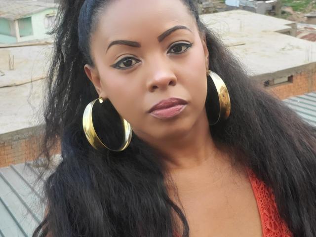 Close-up of RitaClarence. She has medium brown skin, long black hair in a ponytail, and wears large gold hoop earrings. Her eyebrows are thick and arched, and she has black eyeliner with a subtle wing. She wears a red lace top and has pinkish lipstick. The background is blurred, showing rooftops and some greenery. The composition focuses on her face and earrings. She has a neutral expression. The lighting is natural.