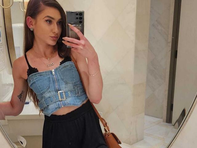 BarbaraLustt with long brown hair, wearing a denim crop top showing her small tits and black skirt, taking a mirror selfie, holding a phone, tattoo on her right arm.