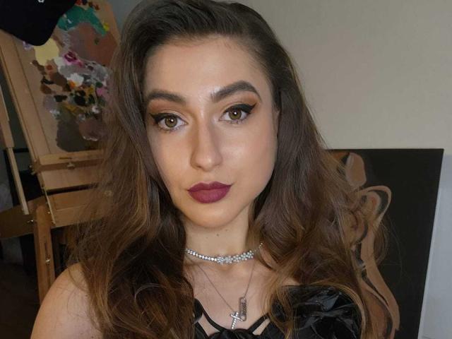 BarbaraLustt heeft lang, bruin, golvend haar, donkerrode lippenstift en een zilveren ketting met een kruis. Ze draagt een zwarte top met spaghettibandjes en kijkt naar de camera..