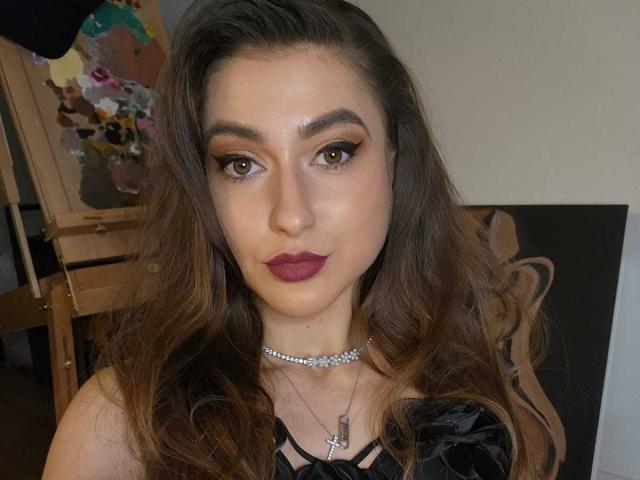 BarbaraLustt with long brown hair, dark lipstick, and smokey eye makeup, wearing a black strappy top and a sparkly choker, looking directly at the camera.