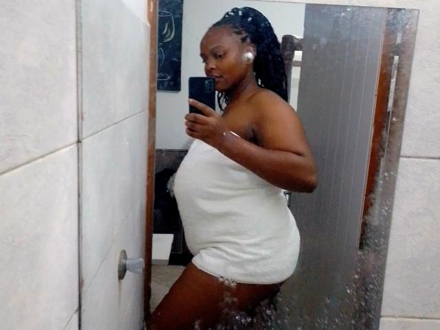 Tinna-hot with dark skin, black braids, and white towel covering her ass and tits, holding a phone in a mirror selfie. She's wearing white earbuds and a bracelet.