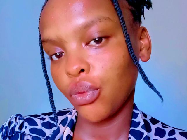 Tinna-hot's face with dark braided hair, brown eyes, and slightly puffed lips, wearing a blue and white patterned shirt, has a smooth, shiny brown complexion.