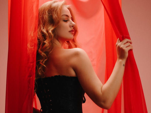 AngyyEyess with curly red hair, wearing a black lace-up corset, holding red curtains, looking over shoulder, soft light, elegant, sexy, subtle tattoo on right wrist.