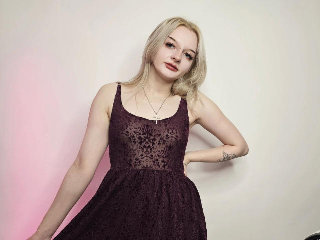 AlessaKiss: atractiva, con piel pálida, cabello rubio y lápiz labial rojo, vestida con un vestido de encaje oscuro y translúcido color púrpura, con las manos en las caderas, un tatuaje pequeño en el brazo izquierdo, mirando a la cámara..