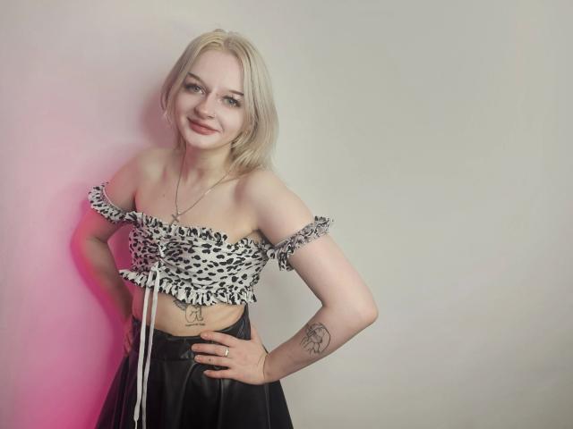 AlessaKiss - aantrekkelijk met blond haar, top met luipaardprint en open schouders, zwarte rok, tatoeage op de rechterarm, lachend, handen op de heupen, roze en witte verlichting. Borsten, billen, vagina, borsten niet zichtbaar..