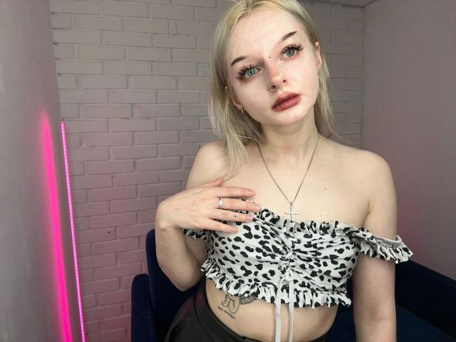 AlessaKiss-hot, a young woman with fair skin and blonde hair, stands in a room. She has blue eyes, a nose ring, and wears long eyelashes. She's dressed in a leopard-print, off-shoulder crop top with ruffled edges, revealing a tattoo on her right side. She's holding her left hand to her chest. Her right hand rests on a dark blue couch. A pink neon light is on the left. Her expression is neutral. The image is sharp with natural light.
