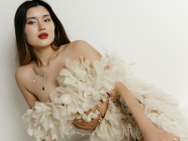 AnneTLuis with bare tits, covered by fluffy white petals, wearing a necklace with green gem, tattoo on right arm, brown hair, light skin, red lipstick, white background.