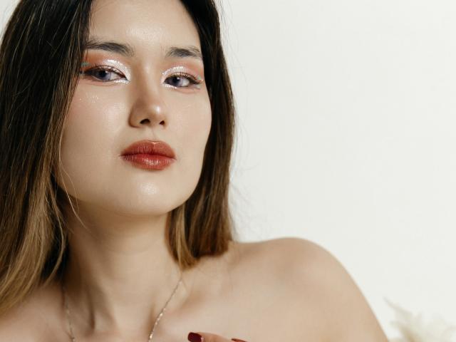AnneTLuis has shiny skin, glossy pink lips, and brown eyes with glittery orange eyeshadow. She's topless, with long brown hair, and a silver necklace.