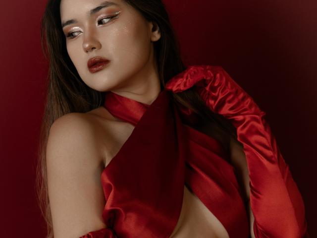 AnneTLuis with glossy red lips, wearing a shiny red halter top and matching red satin gloves, exposing her bare tits. She has long brown hair and sultry makeup.