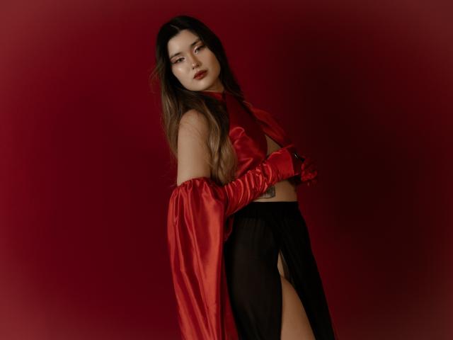 AnneTLuis with long brown hair, red satin cape, and gloves, black sheer pants, showing her tits, standing against a red background, looking seductively at the camera.