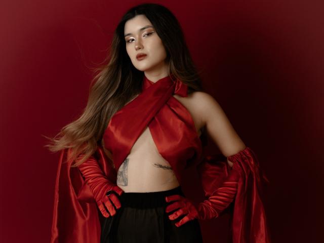 AnneTLuis has long brown hair, red satin wrap dress, exposing her tits, black skirt, red gloves, tattoo on her side, and a confident expression. She's standing against a red background.