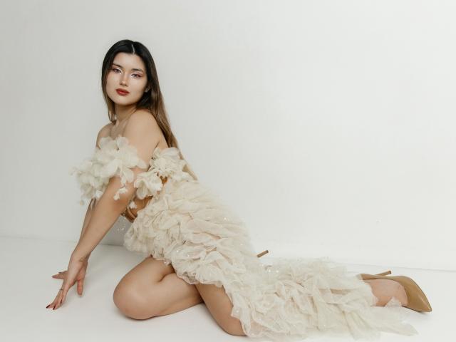 AnneTLuis, Asian woman, kneeling on white floor, wearing sheer, white, ruffled dress, exposing her tits, with beige high heels, long brown hair, red lipstick, looking at camera.