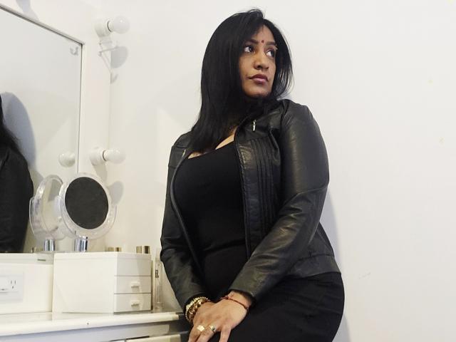 Nahirr with black hair, wearing a black leather jacket and tight black dress, sitting on a white vanity with round mirror, looking to the side, showing her ass.