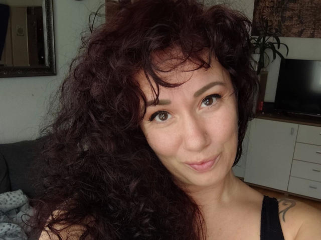 Close-up portrait of Vivien-Molinas with curly, dark red hair covering one side of her face. She has light skin, brown eyes, and is wearing black eyeliner. A small nose ring is in her right nostril. She has a subtle, confident smile. Vivien-Molinas is wearing a black tank top, and a tattoo is visible on her right shoulder. The background is blurred, with a TV and a plant. The image is well-lit, highlighting her facial features and hair texture.