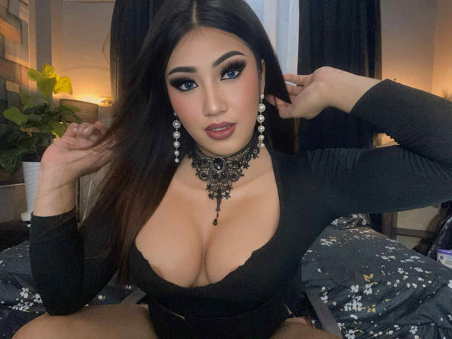 BeckyMcTracy, com seios grandes, um colar de renda preta e brincos longos, sentada em uma cama, vestindo uma blusa preta, com a mão no cabelo, olhando para a câmera..