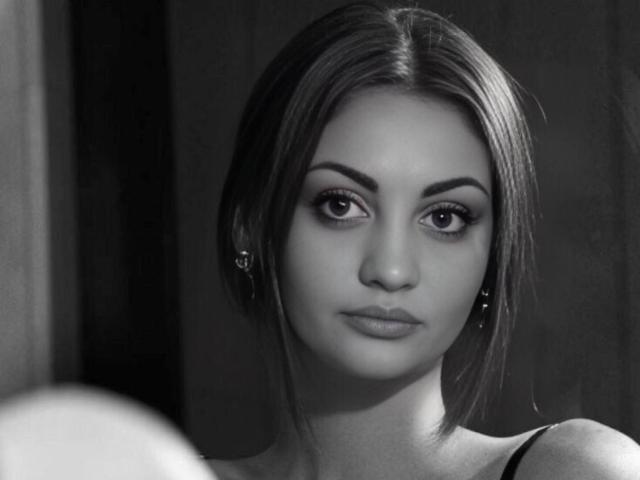 Photo en noir et blanc de TinaLoversX, avec des cheveux lisses de longueur moyenne, portant des boucles d'oreilles, regardant directement l'appareil photo avec une expression neutre, montrant subtilement ses seins dans un haut sans manches..