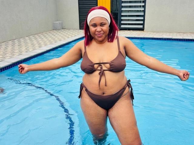 CharmainPhilly69 with red hair, brown bikini, and white headband, standing in a blue pool, arms out, water covering her round tits and ass, smiling.