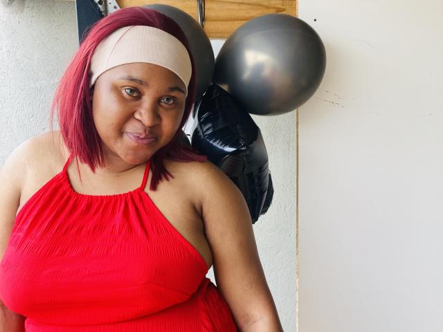 CharmainPhilly69 with red hair, beige headband, and red dress, showing her tits, smiling, gray and black balloons behind her, white wall. She's medium brown, slightly chubby.