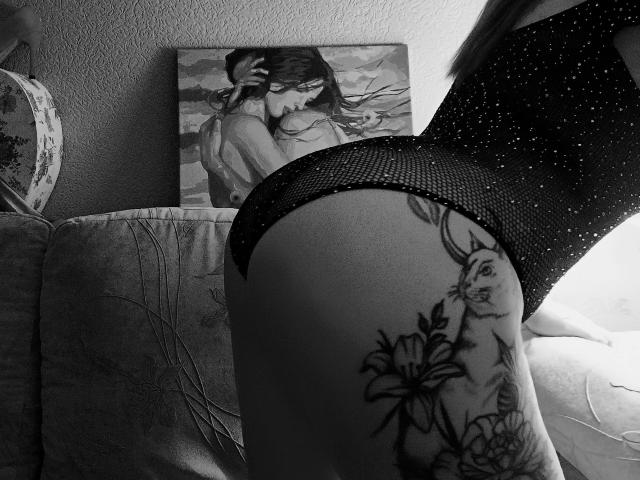 Niferttiti in a sparkly black mesh bodysuit, showing a tattooed ass with a bunny and flowers, leaning on a couch, black and white photo, her back to camera.